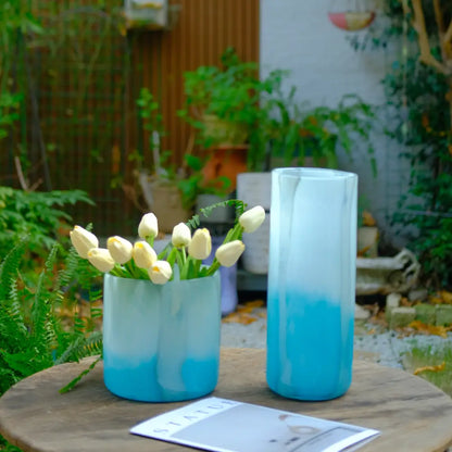 Design vase en verre artisanal avec effet ombré bleu, associé à un bouquet de tulipes claires.