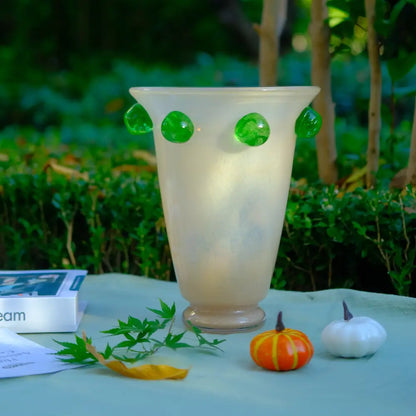Grand vase en verre crème orné de pastilles vertes, posé en extérieur dans un décor naturel.