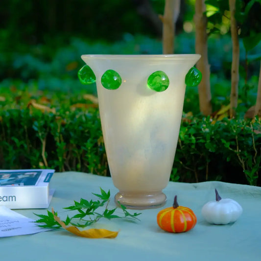 Grand vase en verre crème orné de pastilles vertes, posé en extérieur dans un décor naturel.