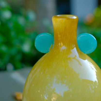 Vase jaune en verre soufflé avec deux anses rondes bleues, au design moderne et raffiné.