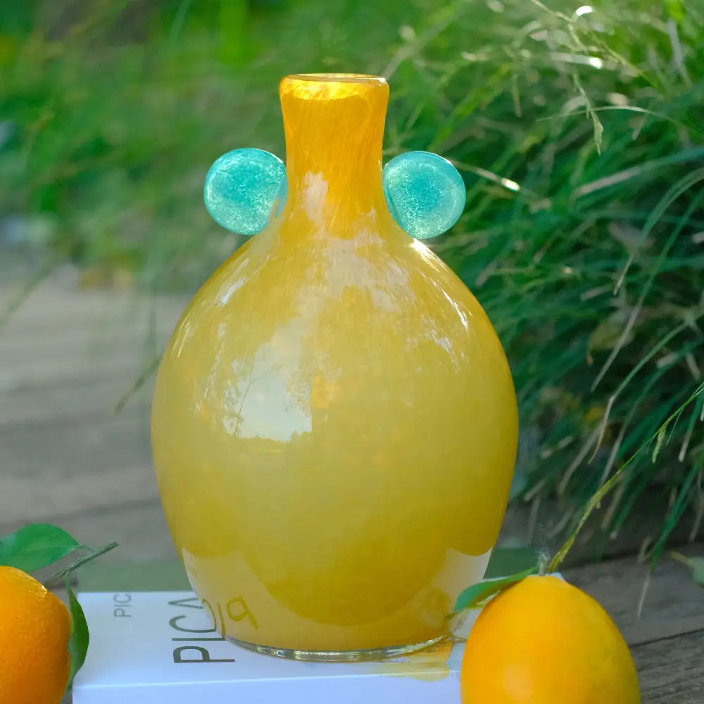 Vase jaune en verre soufflé rond, orné de deux disques verts, posé dans un décor naturel.