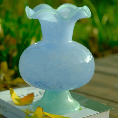 Vase artisanal en verre soufflé bleu pâle avec col ondulé et pied élégant, photographié en extérieur.
